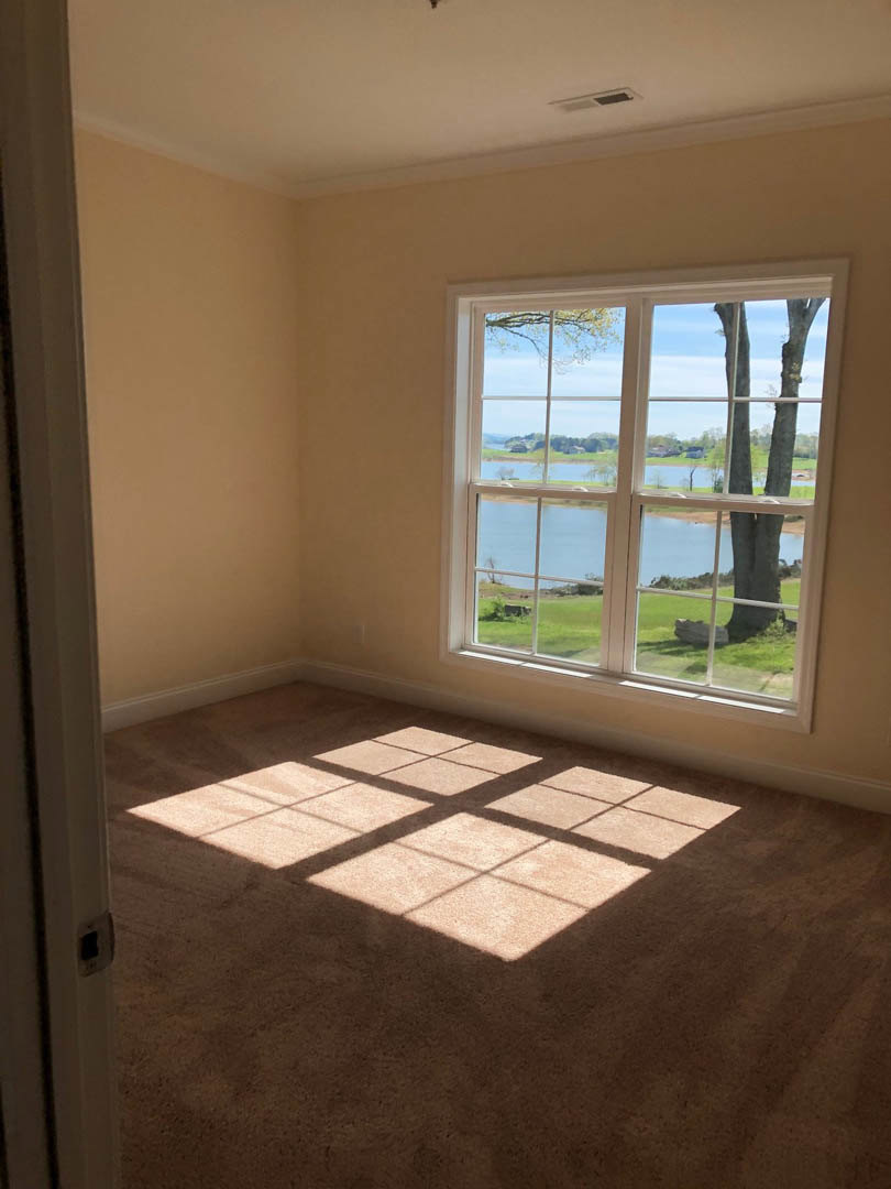 Sunlit room with large window overlooking lake and grassy shoreline, square patterned floor, white walls, and partial view of door