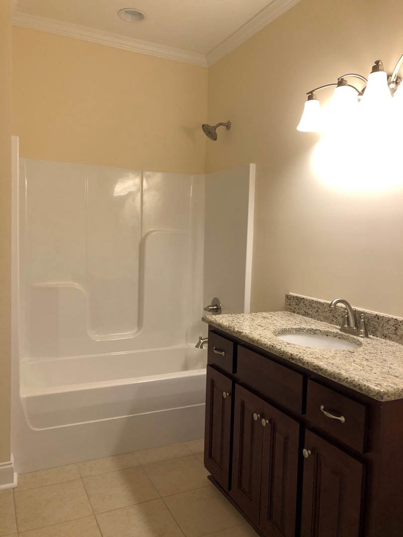 Granite countertop vanity with undermount sink, three-light fixture above, tiled shower with wall-mounted showerhead, white bathtub, stone accent surfaces, and wood cabinetry.