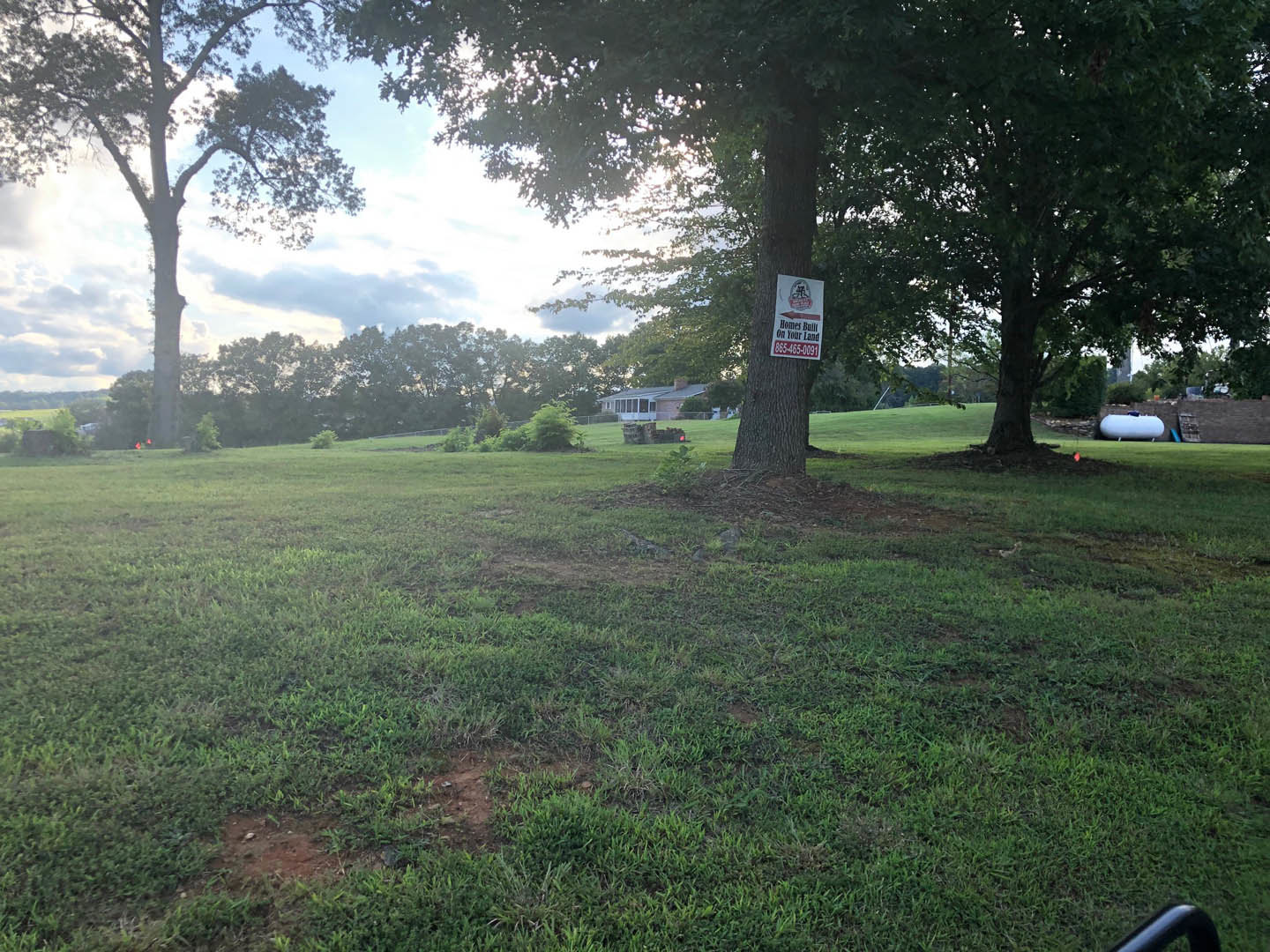 Tree with a white sign attached, surrounded by green grass and open lawn under a partly cloudy sky