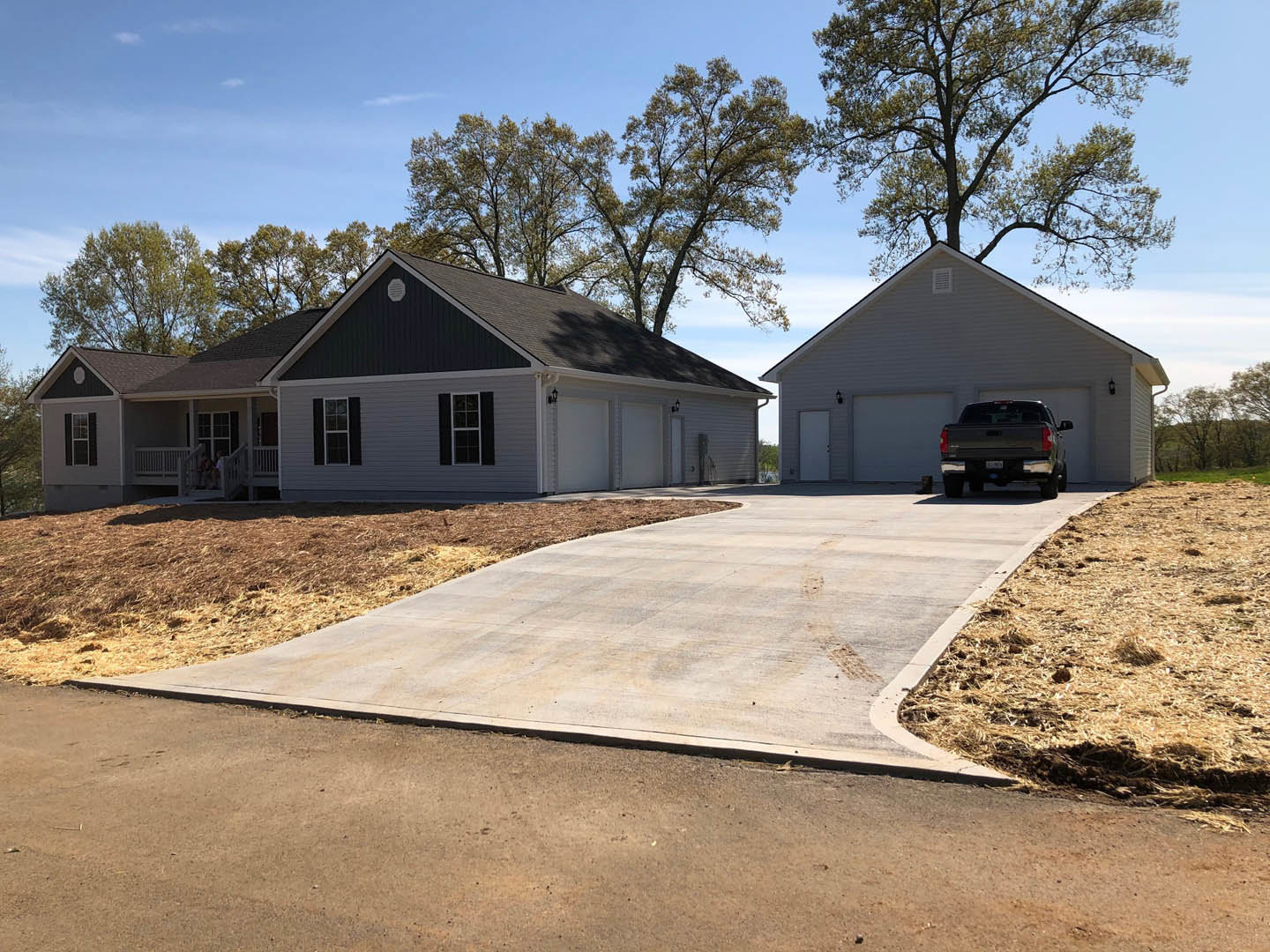 Two-story home with gray siding, attached garage, concrete driveway, black pickup truck parked in front, small tree and lawn, person walking past large front window