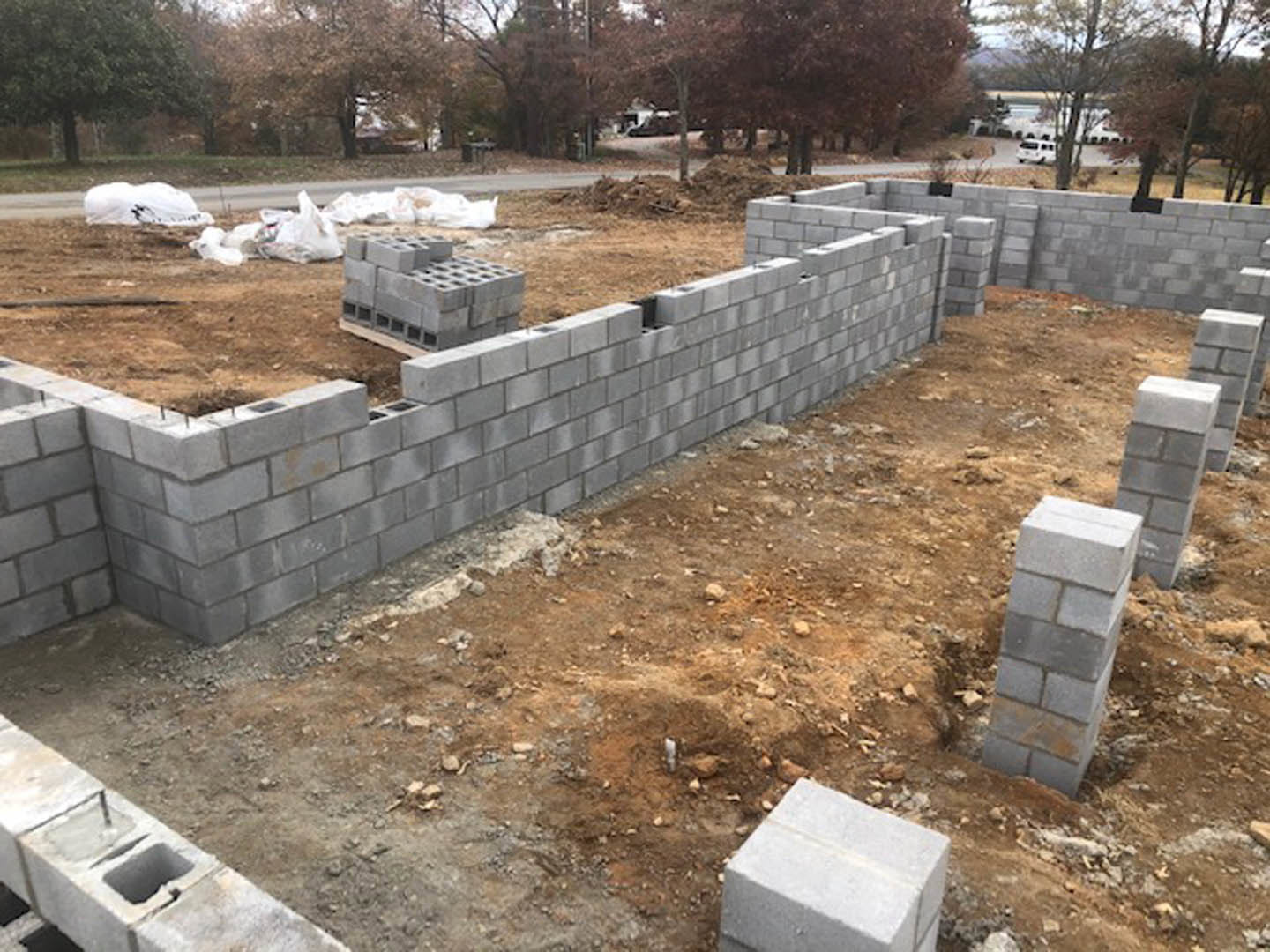 Concrete foundation blocks and brick pillars set on dirt ground at a residential construction site, with a partially built grey brick wall and scattered white blocks.