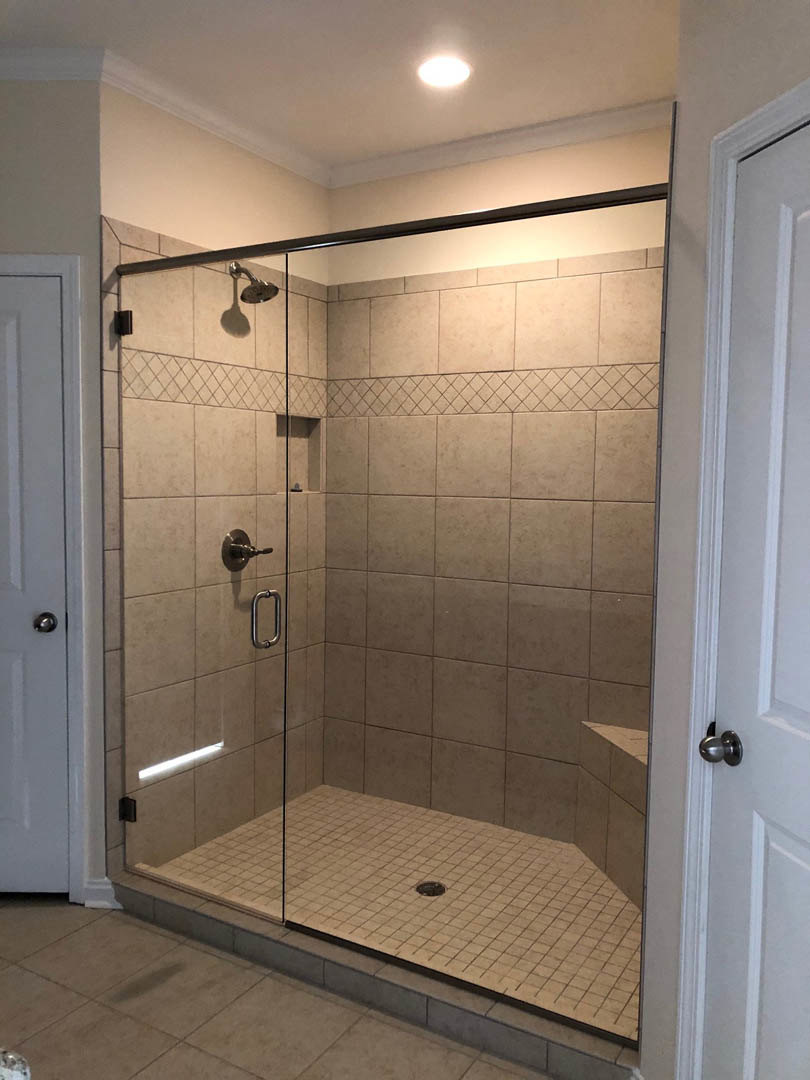 Glass shower enclosure with tile floor, built-in bench, wall-mounted plumbing fixtures, and a door featuring a metal handle; mirror reflects bathroom interior.