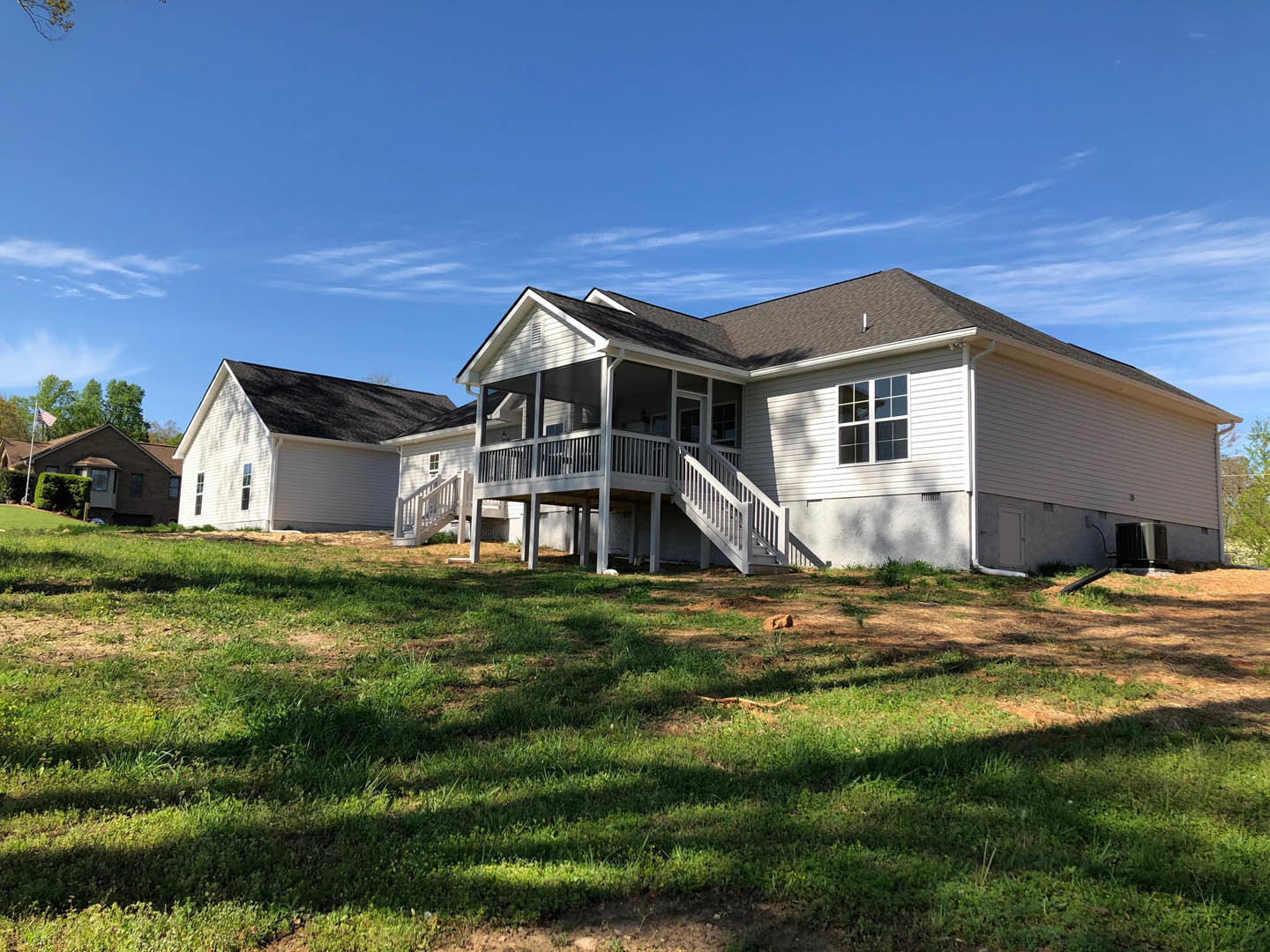 White house with wraparound porch on grassy hill, American flag near entry, trimmed hedge along front, Southfork Ranch visible in background under partly cloudy sky