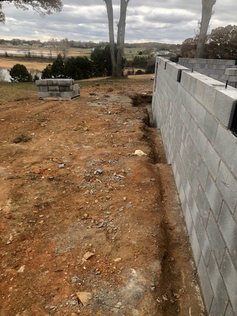 Smooth grey concrete wall bordering a modern building, with tall trees and cloudy sky in the background, soil and rocks at the base