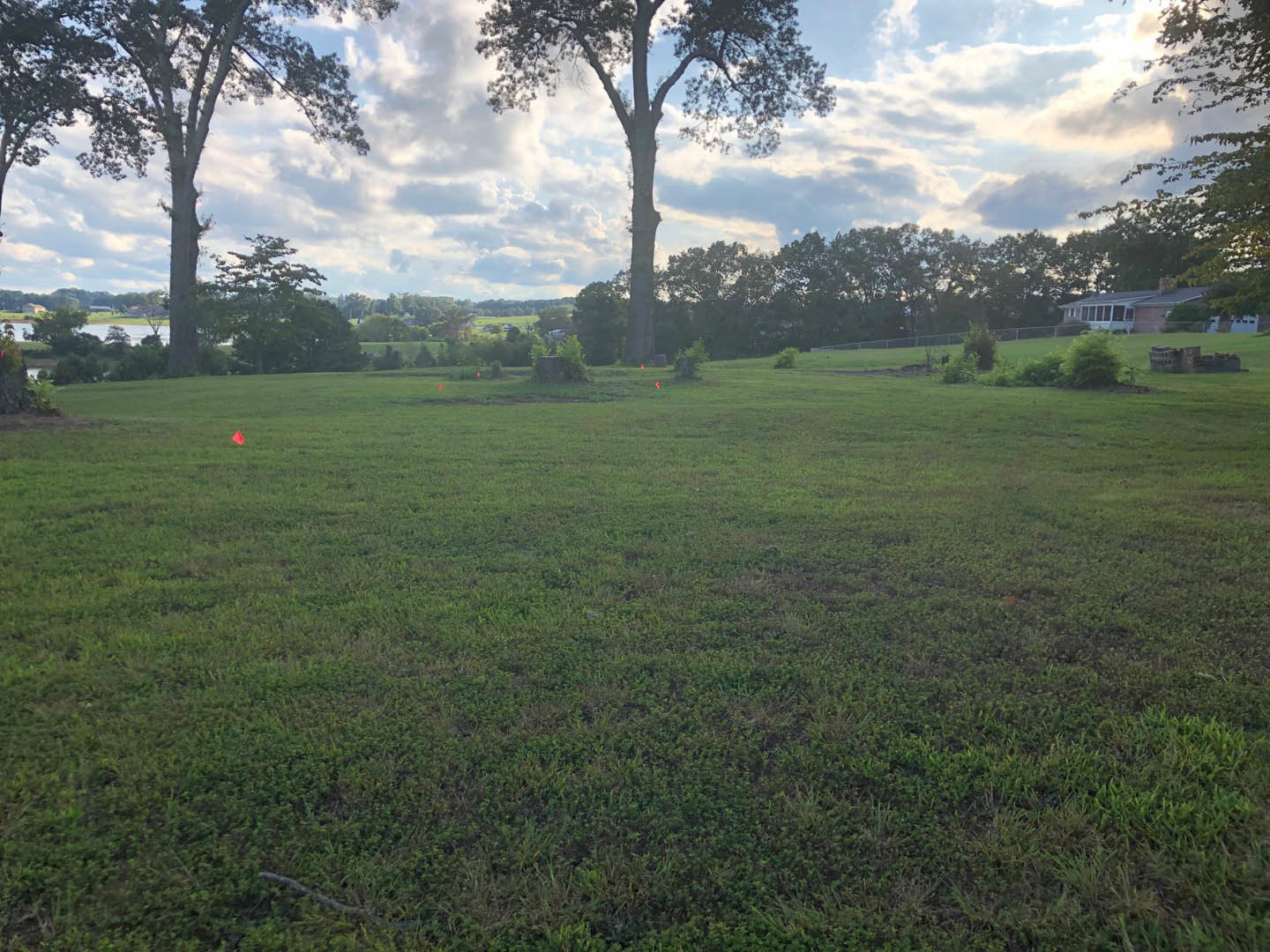 Expansive green lawn bordered by mature trees, orange survey markers scattered across grass, distant house with chimney partially visible, cloudy sky overhead, small pond beyond