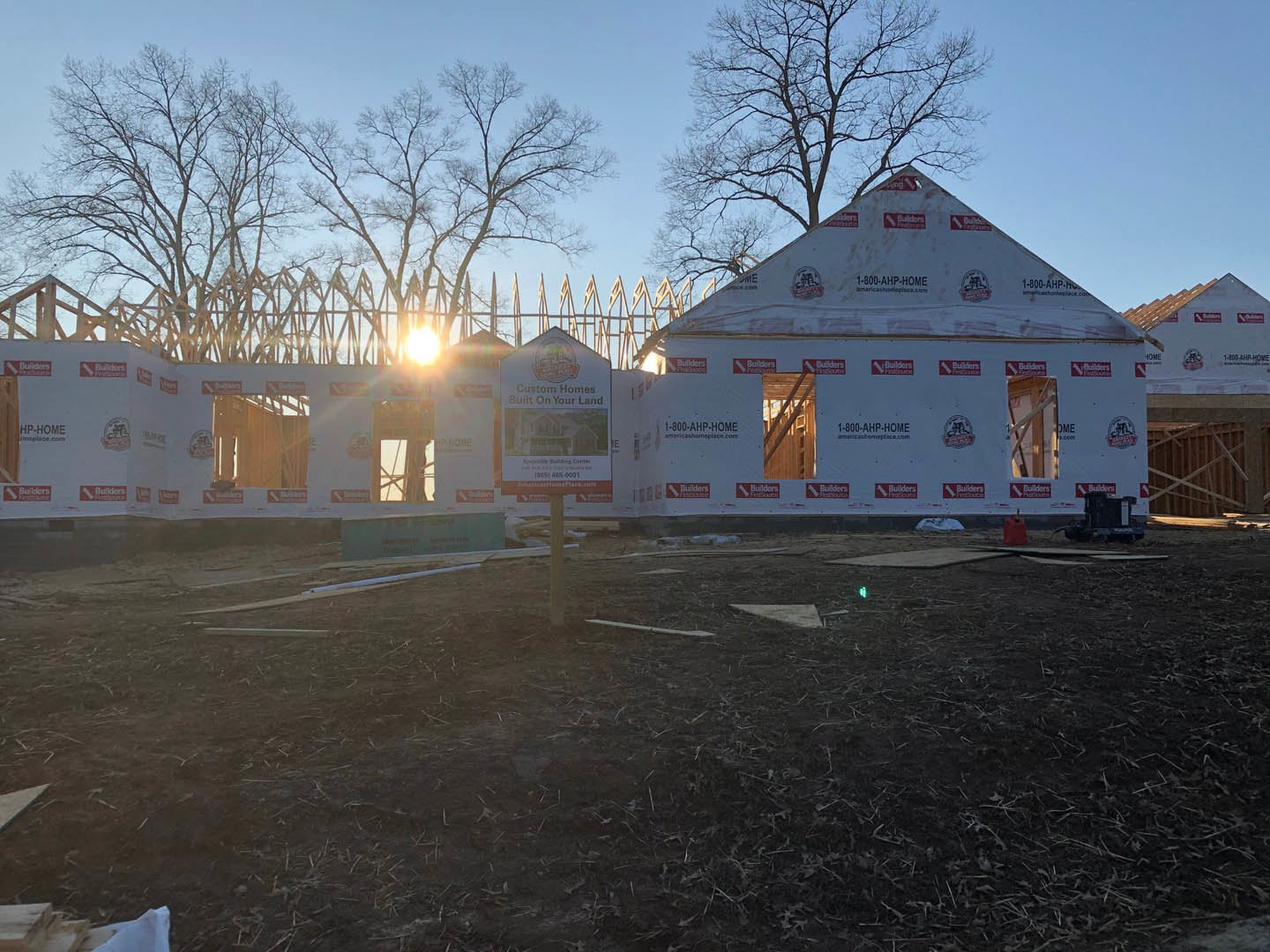 White house under construction with exposed framing, dirt field in foreground, construction sign attached to exterior wall, leaf pile and utility pole nearby, bare trees and