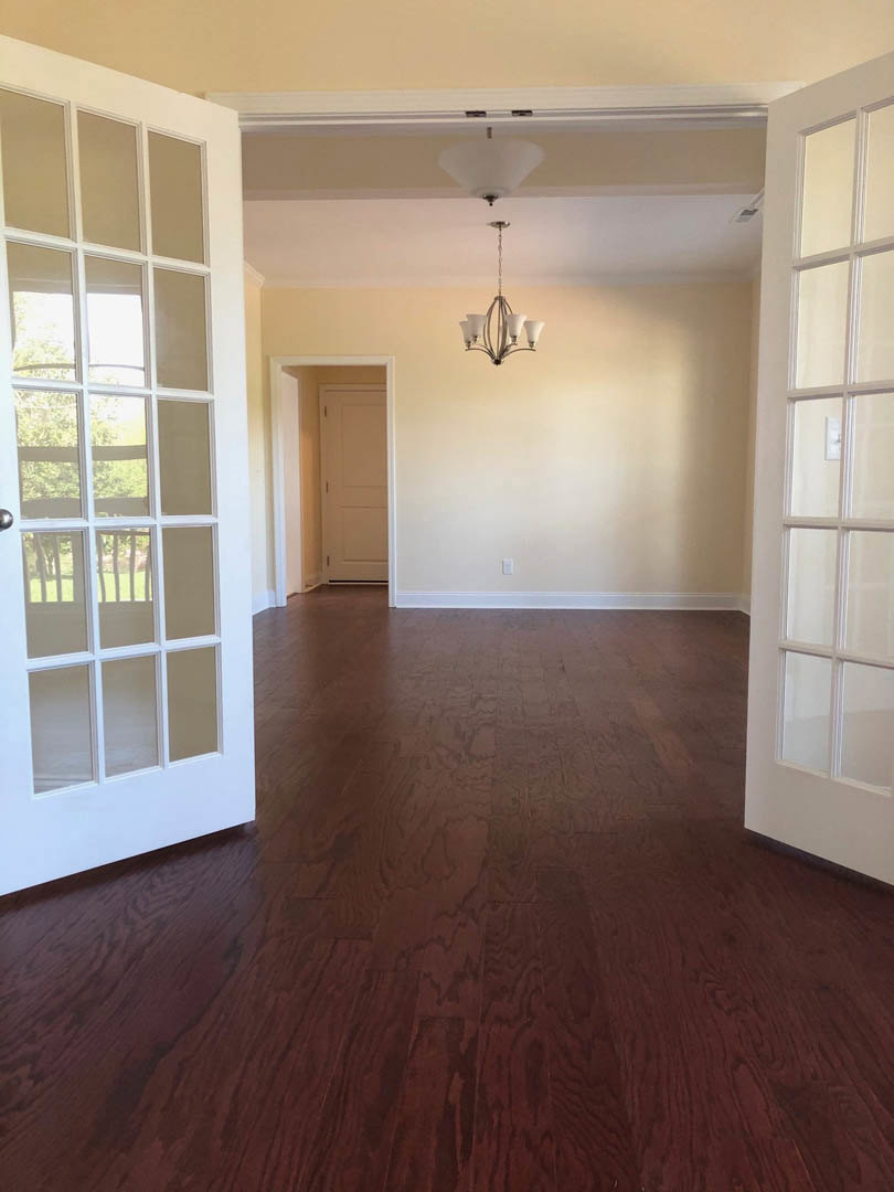 Room with hardwood flooring, white paneled door with metal handle, large window, and ornate chandelier hanging from a white ceiling.