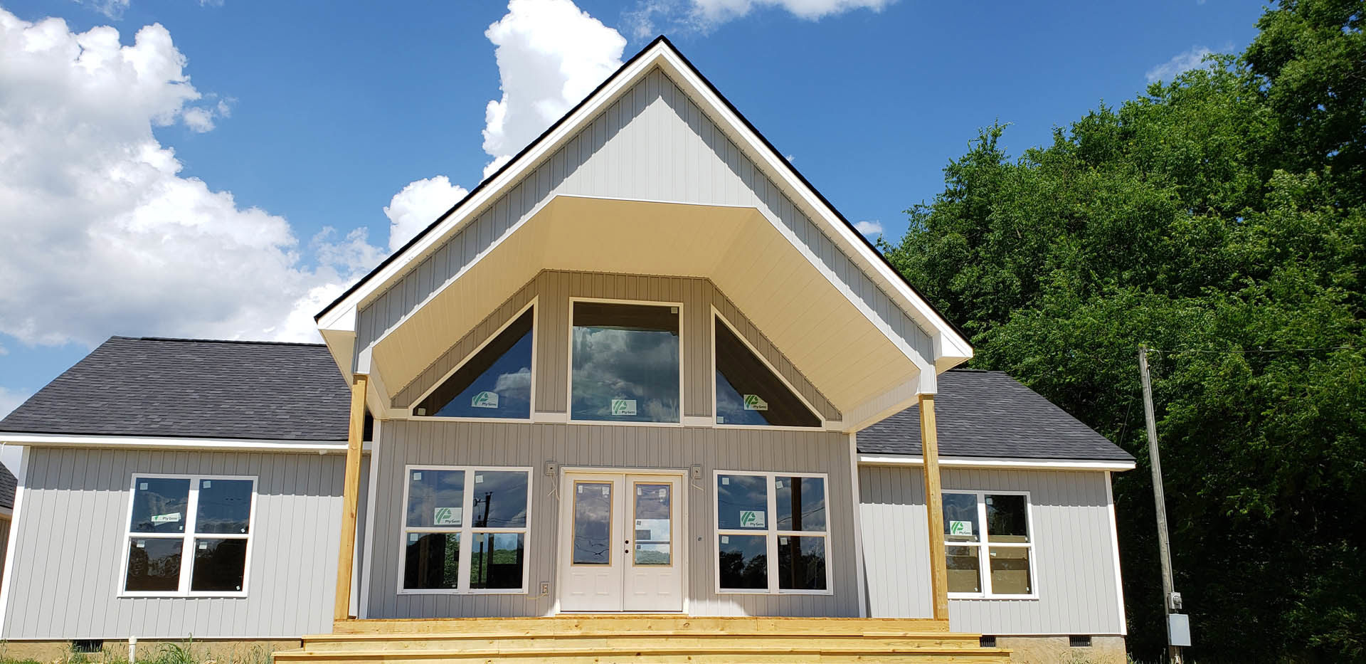 Two-story custom home under construction with gray siding, white-framed windows, double entry doors, covered porch, and surrounding trees beneath a blue sky