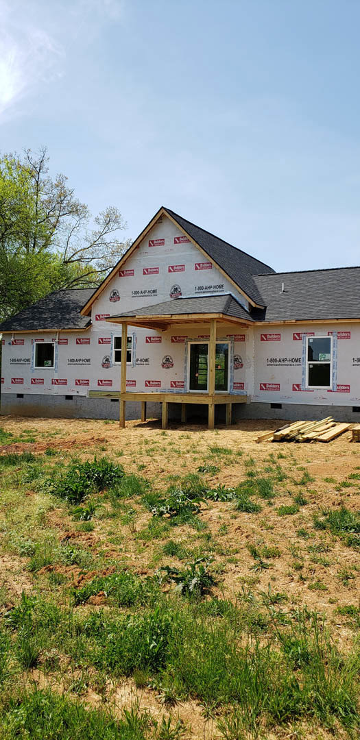 Partially built house with exposed wooden beams, double glass-panel doors, two porches, pile of lumber on dirt and grass, window displaying a sign, cloudy sky overhead