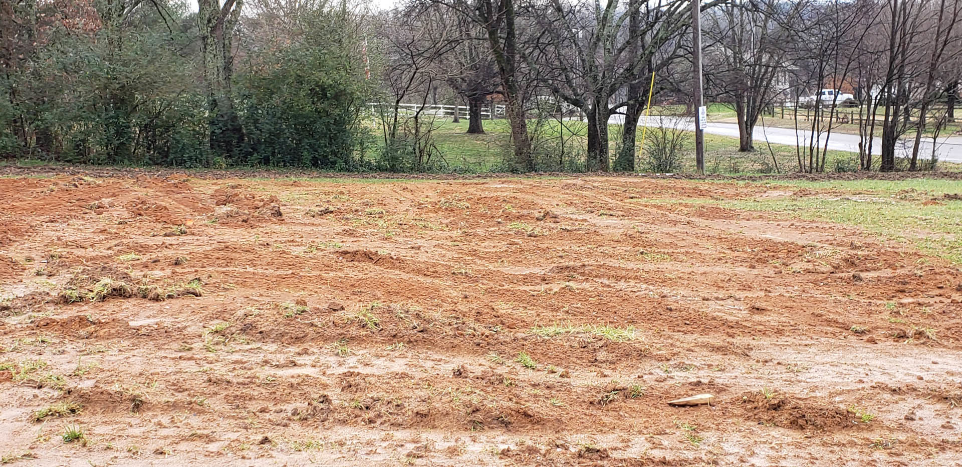 Dirt field with patches of grass, group of trees in the background, road along the edge, clear sky overhead