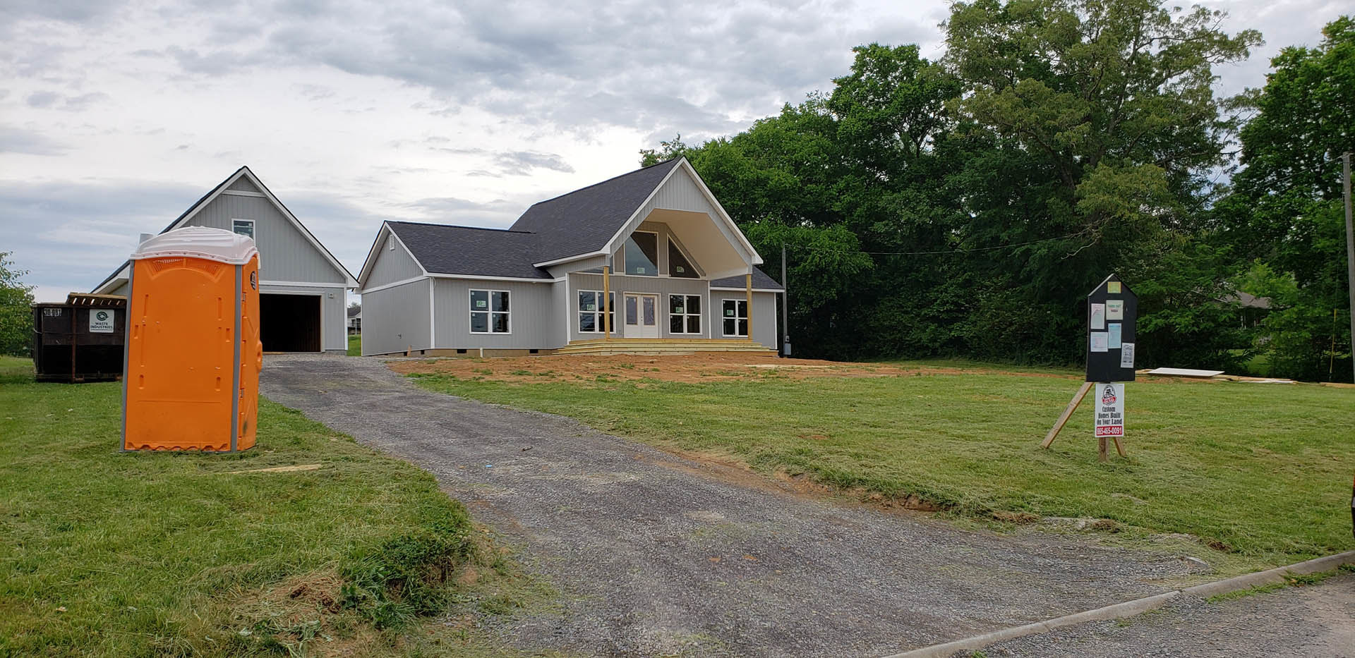 Two-story house with white double doors and glass windows, covered porch, gravel driveway, orange portable toilet, construction sign, surrounded by trees and grass under cloudy sky