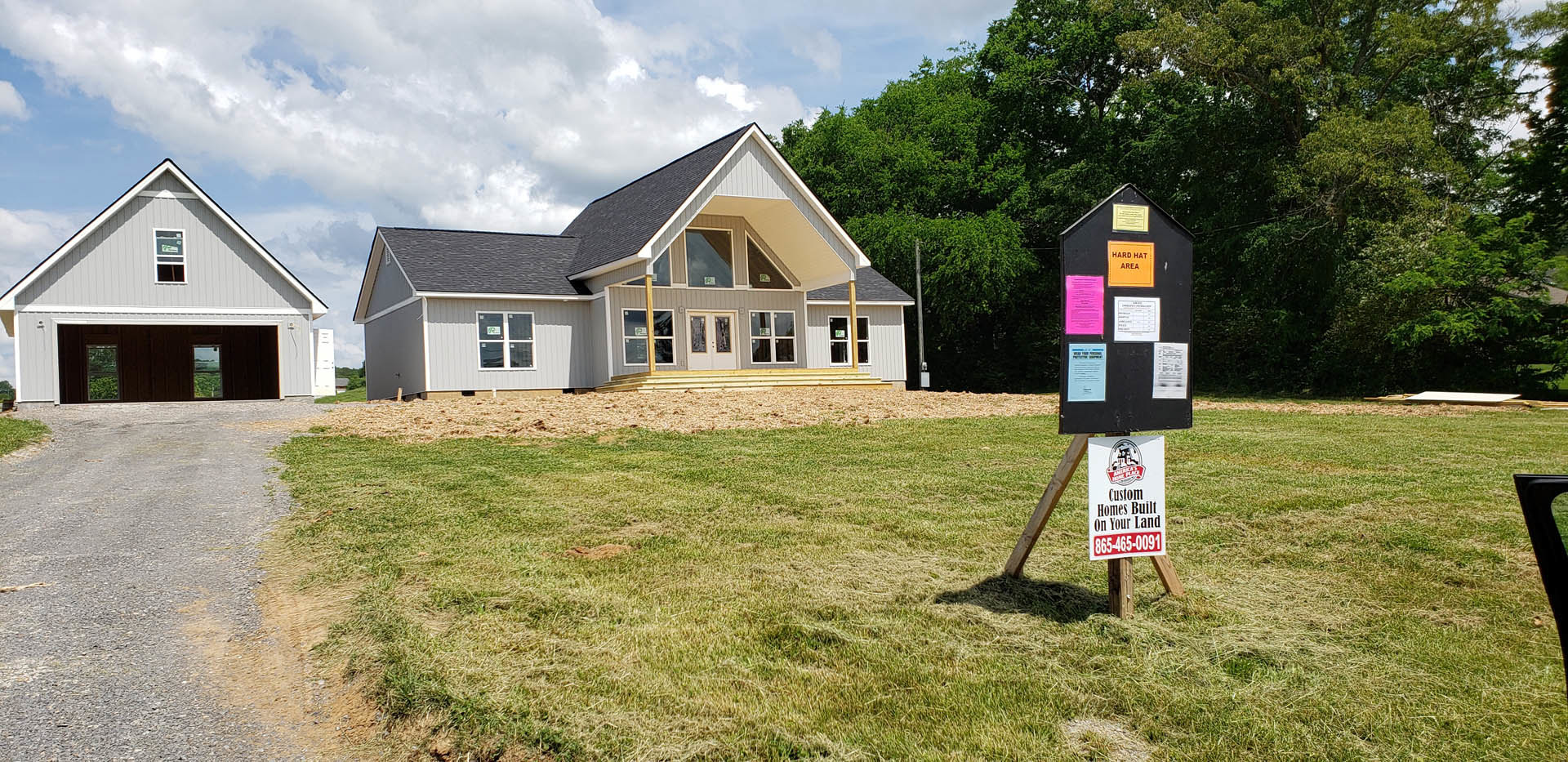 Two-story house with attached garage, white-framed windows, gray siding, yellow sign with black text on a pole in front, cloudy sky overhead, grassy yard.