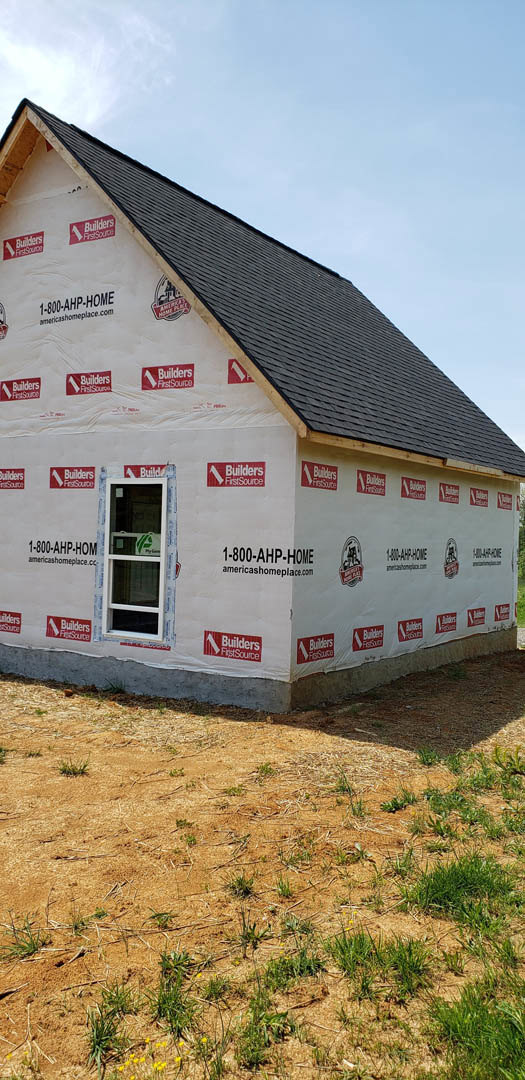 White-wrapped house under construction with red and white logo signage, large window, dirt and grass field in foreground, cloudy sky overhead