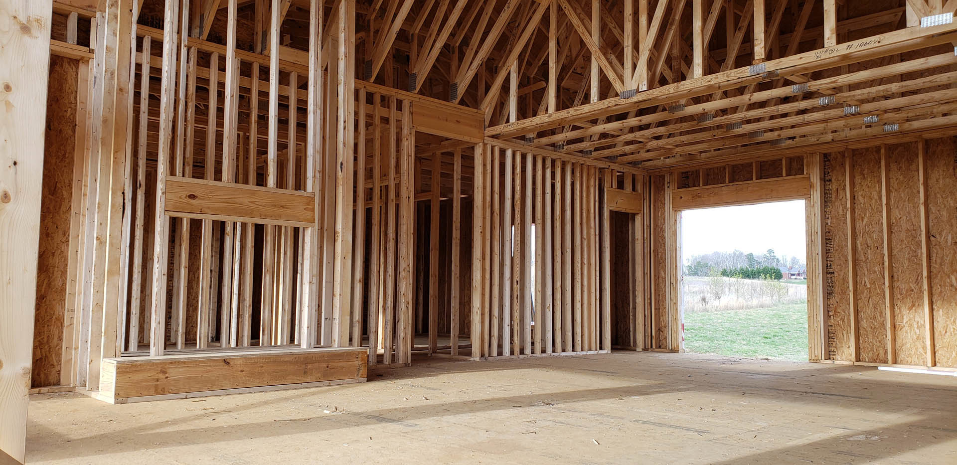 Exposed wooden framing and roof trusses of a house under construction, sunlight casting shadows on the ground, open window framing a view of grassy field, close-up details of