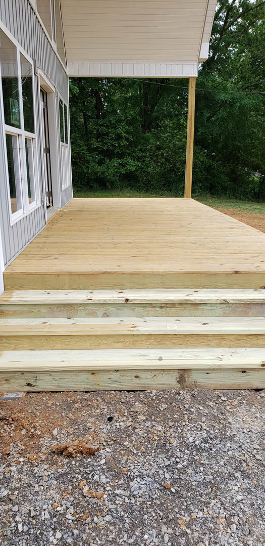 Wooden deck with stairs leading to gravel surface, bordered by white paneled wall and window, overlooking forest through glass.