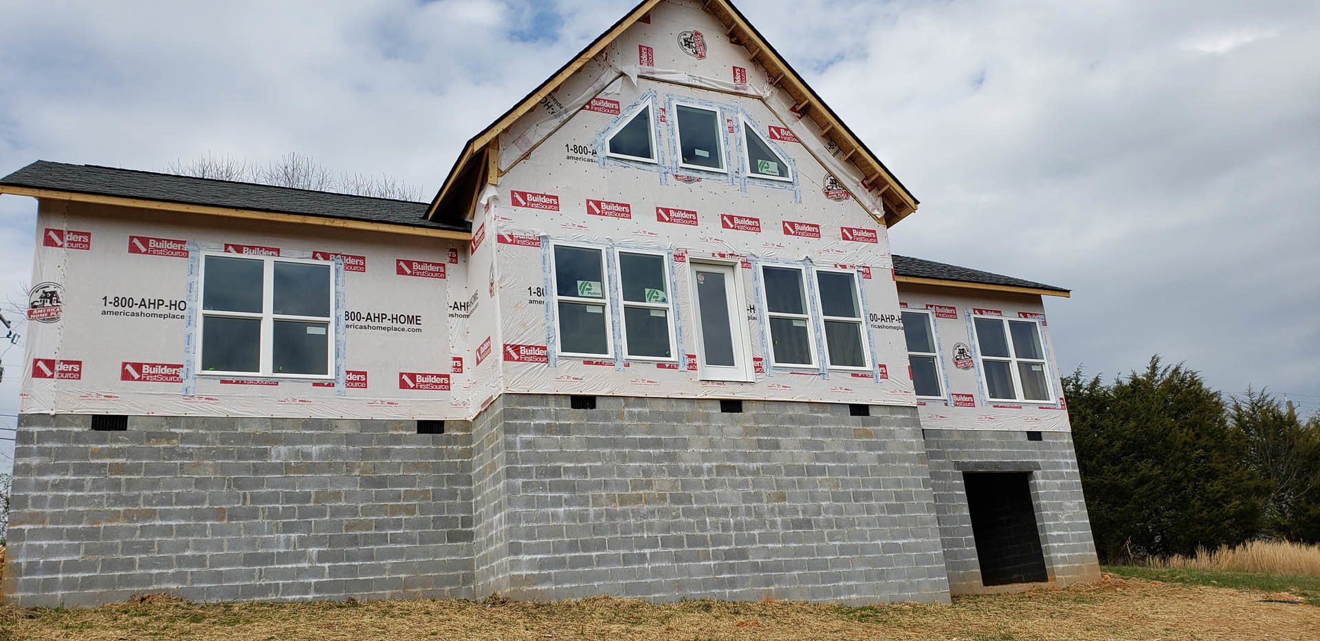 Partially built brick house with red and white adhesive tape on window frames, tall grass in foreground, trees in background, dark doorway visible, window displaying a sign