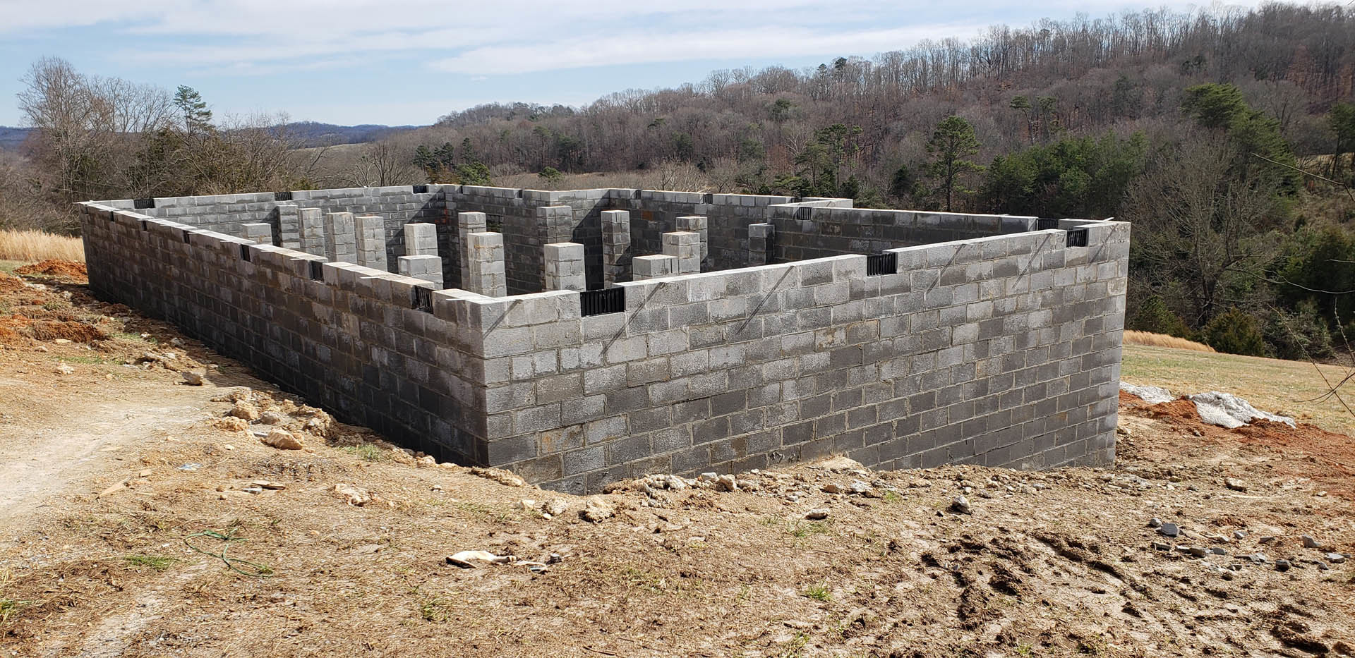 Partially built stone exterior walls surrounded by dirt ground, scattered construction materials, and leafy trees in the background under a cloudy sky