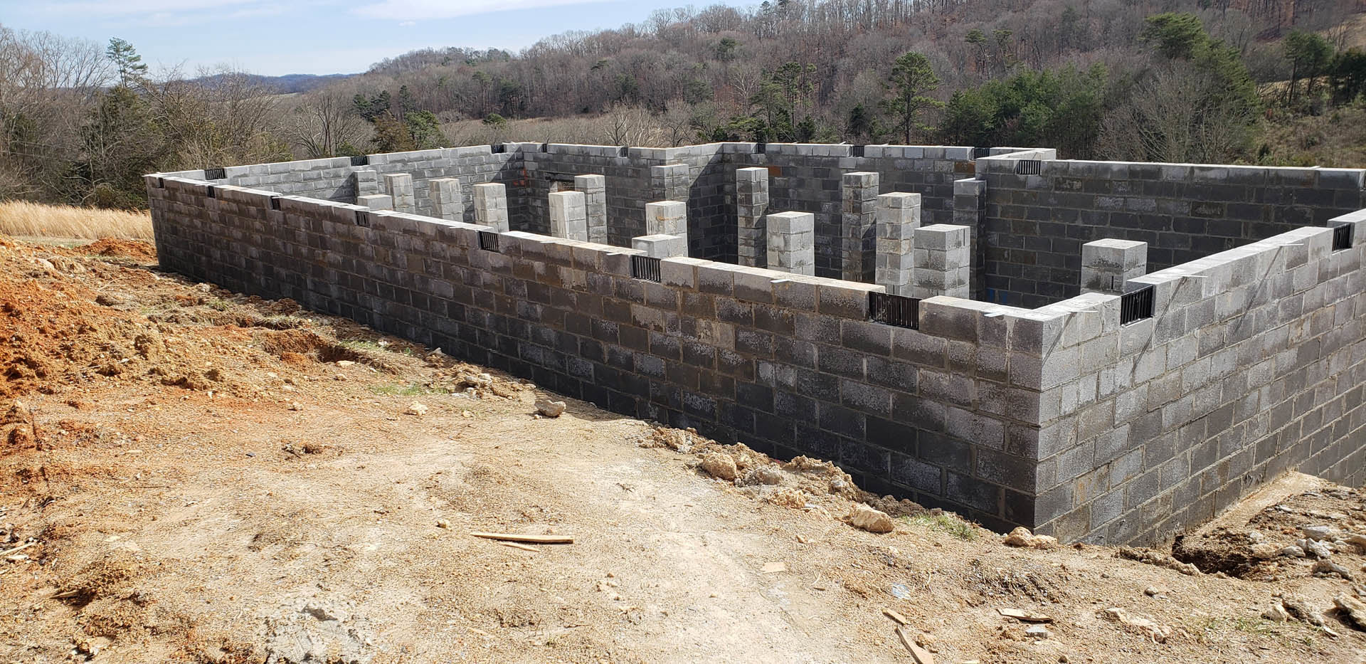 Concrete block wall with vertical columns, black metal fence on top, white cube structure with yellow band nearby, stone wall and trees in background, building site under open sky.