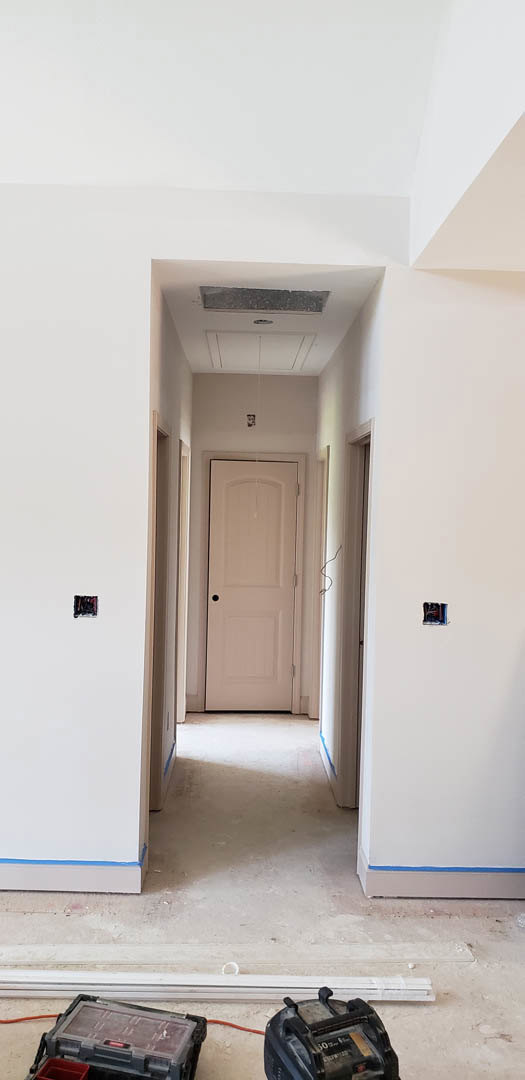 Hallway with light-colored walls, white door featuring a black handle, wood flooring, ceiling fan, and a small plastic box with a red indicator light mounted near the door.