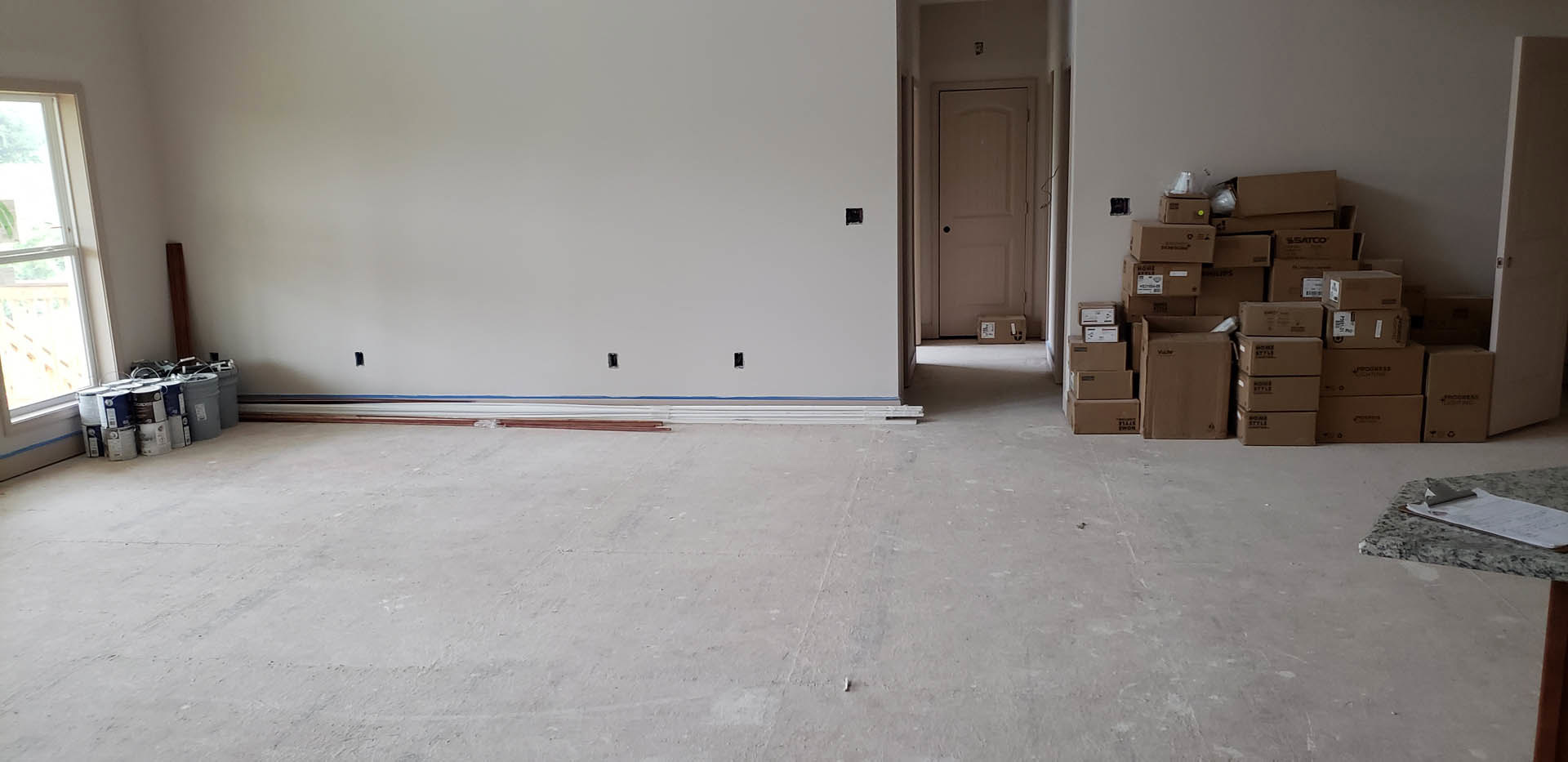 White plaster walls and ceiling, concrete floor, white door with black handle, cardboard moving boxes stacked near wall, window partially visible, empty room with minimal