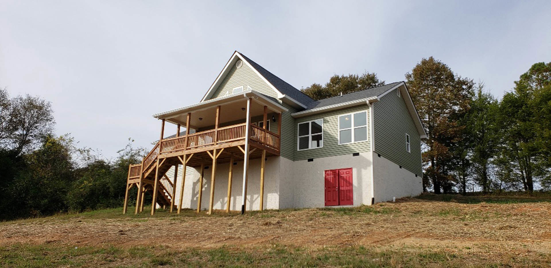 Wooden deck and covered porch on a cottage-style home with red front door, surrounded by grass lawn and mature trees under a clear sky