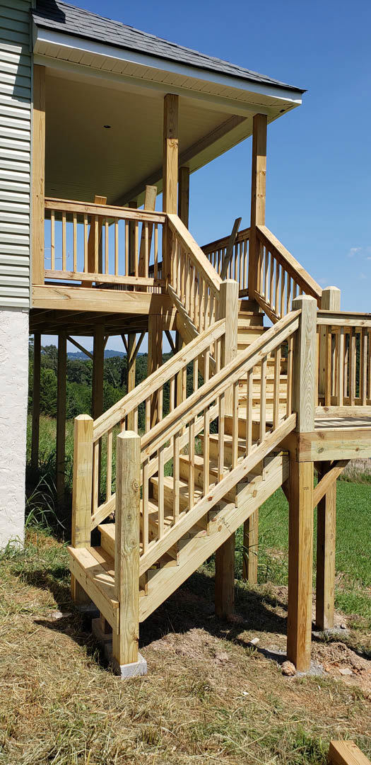 Wooden exterior stairs with handrail leading to covered porch, surrounded by dry grass and plants, gazebo visible in background