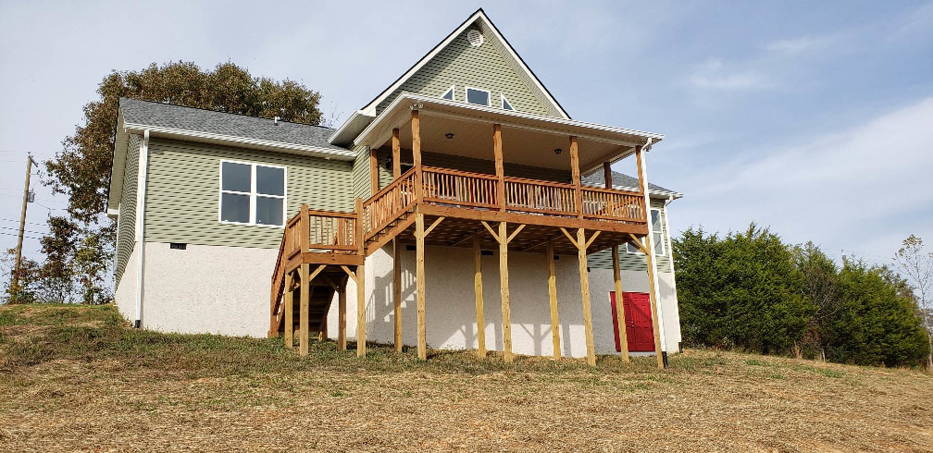 Two-story cottage with wooden deck and covered porch, red front door with yellow stripes, large windows, surrounded by grassy lawn and mature trees under partly cloudy sky