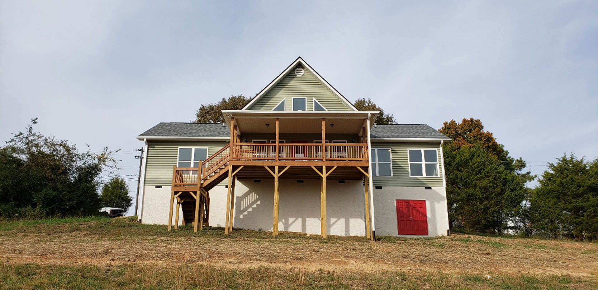 Two-story home with white siding, red front door, covered porch, upper-level deck with railing, manicured lawn, and mature trees in the yard