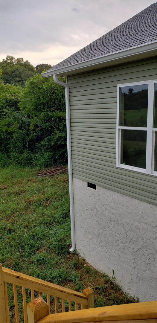 Modern home exterior with large window, gray siding, covered porch, wooden railing, and manicured grass lawn bordered by trees and cloudy sky