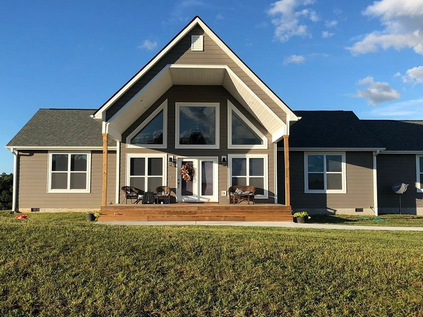 Two-story house with white-trimmed windows, brown siding, covered porch with chairs, manicured green lawn, and wooden fence under partly cloudy sky