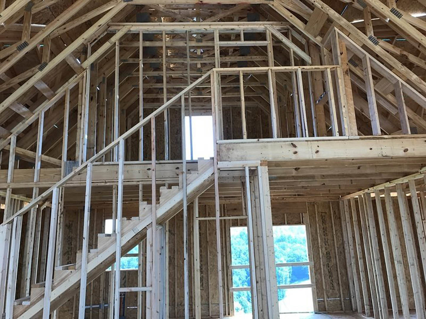 Exposed wood framing and beams inside a house under construction, window opening reveals forest and blue sky outside
