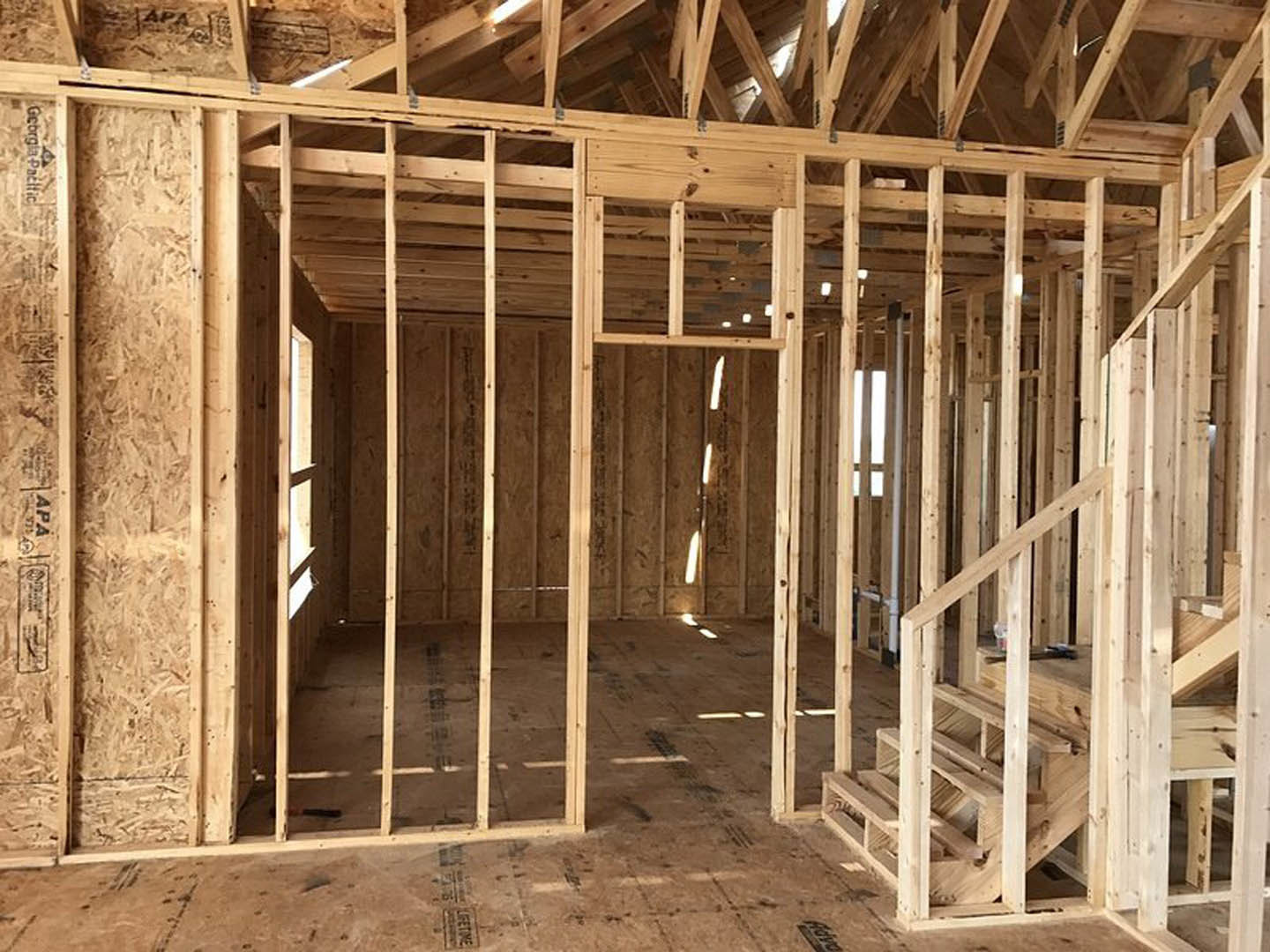 Exposed wooden framing and beams inside a partially constructed house, with visible wall studs, roof structure, and unfinished lumber.