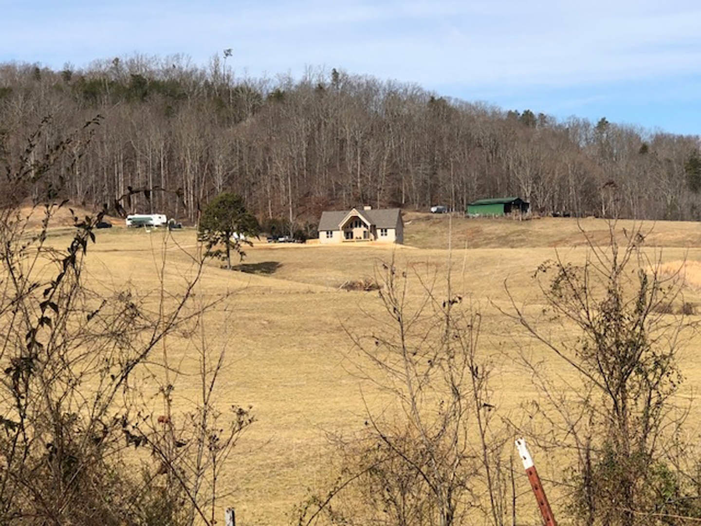 White custom home with dark roof sits on grassy field, surrounded by mature trees and open sky, with a trailer visible in the distance.