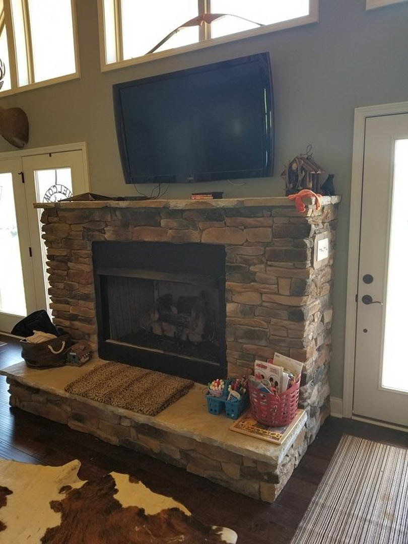Stone fireplace with glass doors, television mounted above, woven basket filled with magazines on hardwood floor, patterned rug in front, white walls and large window in living