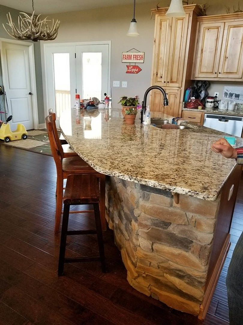Kitchen with stone-faced island, wood cabinetry, floral upholstered chair, potted plant on table, yellow toy car on hardwood floor, antlers displayed nearby