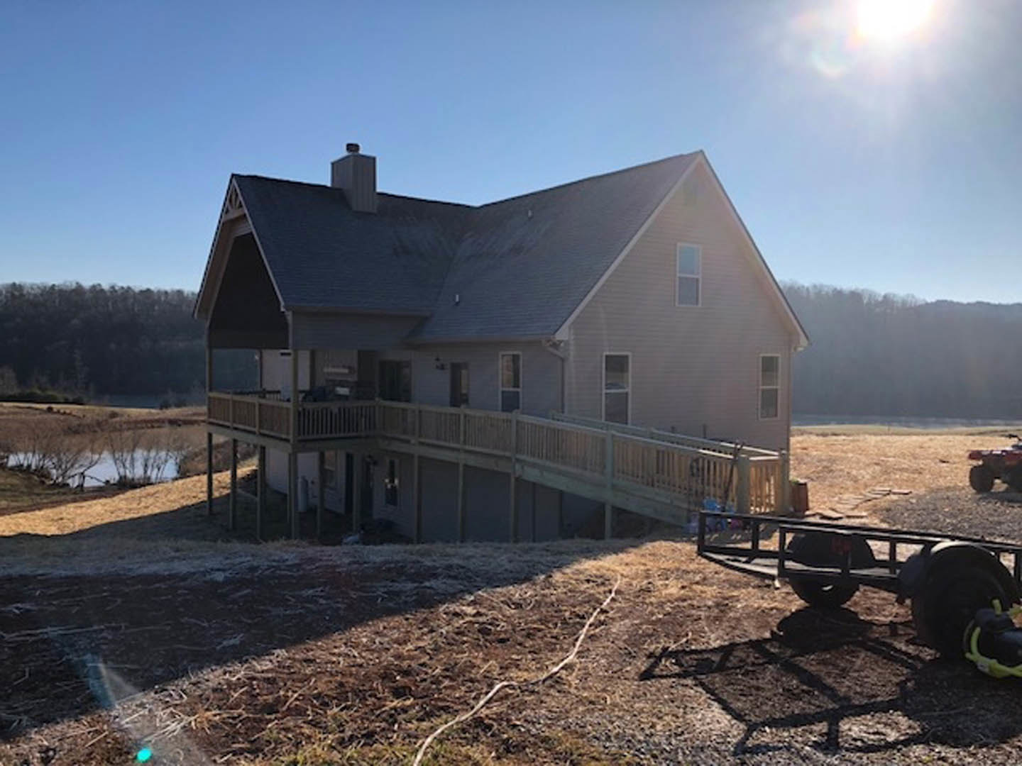 Two-story house with wood deck and upper balcony, white siding, grassy yard, metal-frame trailer parked in front, leafless trees, winter sky