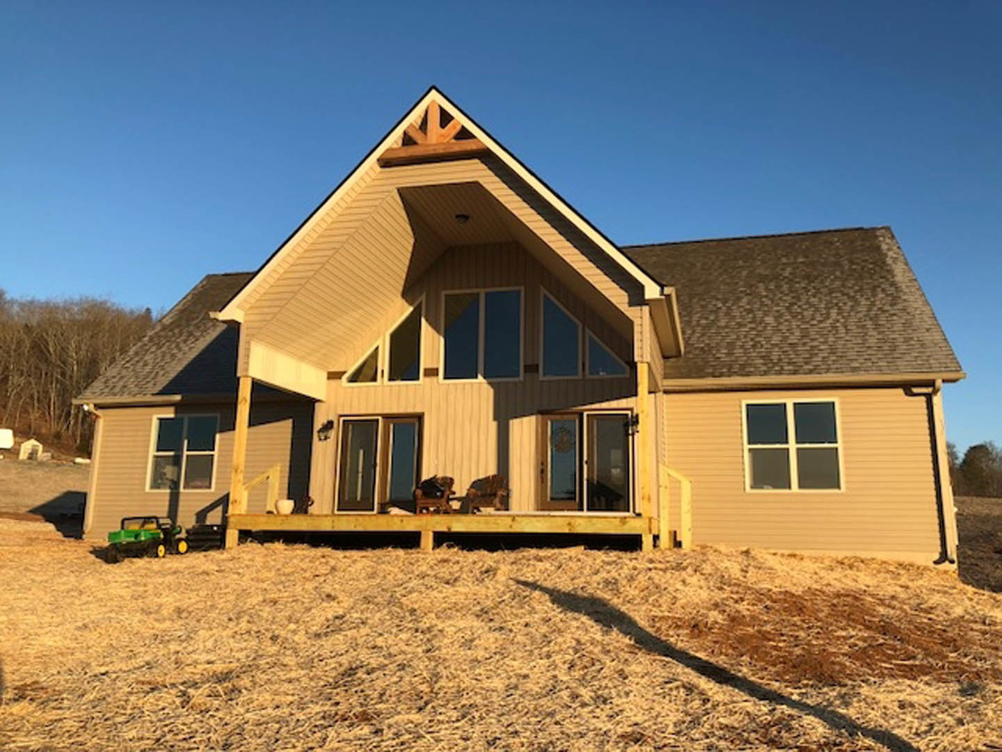 Two-story house with light siding, wooden deck, and porch overlooking dry grass lawn; green and black lawnmower parked near blue sign, large windows visible.