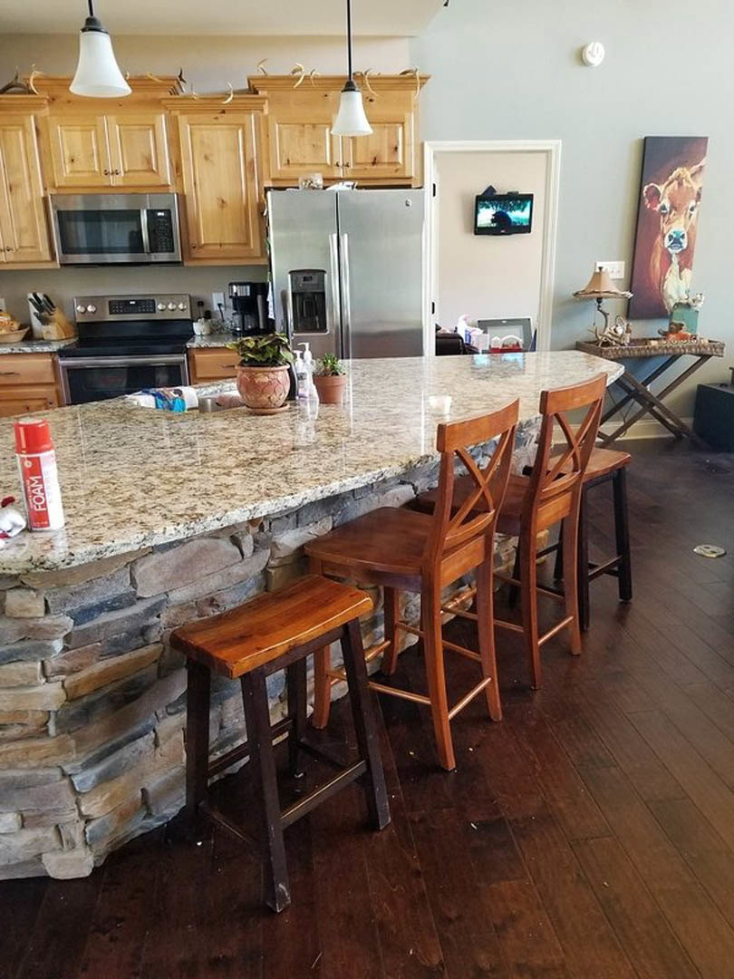 Kitchen with a stone island, stainless steel refrigerator, wood cabinetry, built-in microwave, stove, wooden stool beside a brick accent wall, and bottle on the countertop