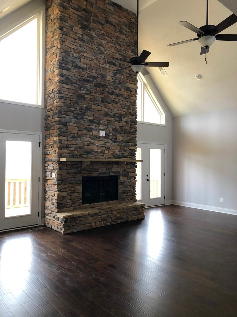 Stone fireplace with wooden mantel, hardwood floor, black door set in brick accent wall, ceiling fan with light fixture, sunlight streaming through doorway