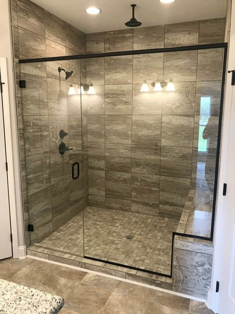 Walk-in shower with clear glass doors, white and black speckled tile walls, black round light fixture, and tile flooring