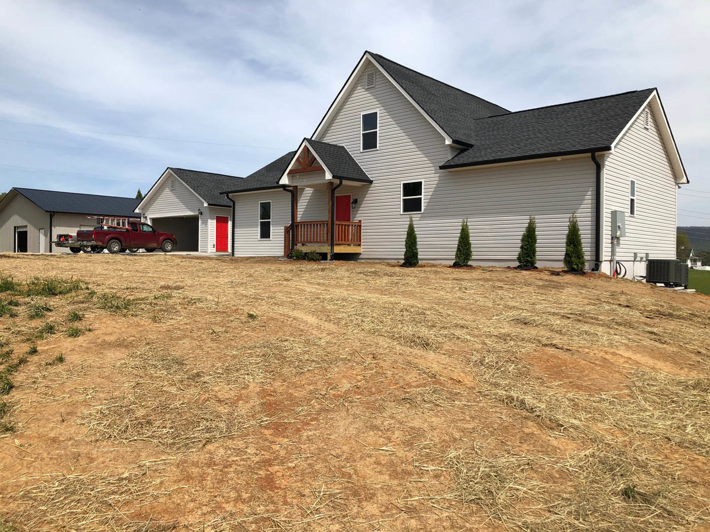 White farmhouse with red front door, red car parked on gravel driveway, grassy yard, tree near white exterior wall, cloudy sky overhead