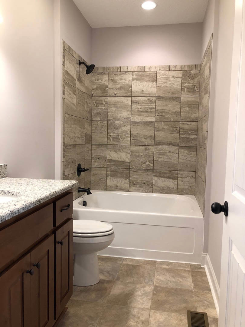 White bathroom with closed toilet, white bathtub, granite countertop, tile floor, black door knob, and black wall fixture