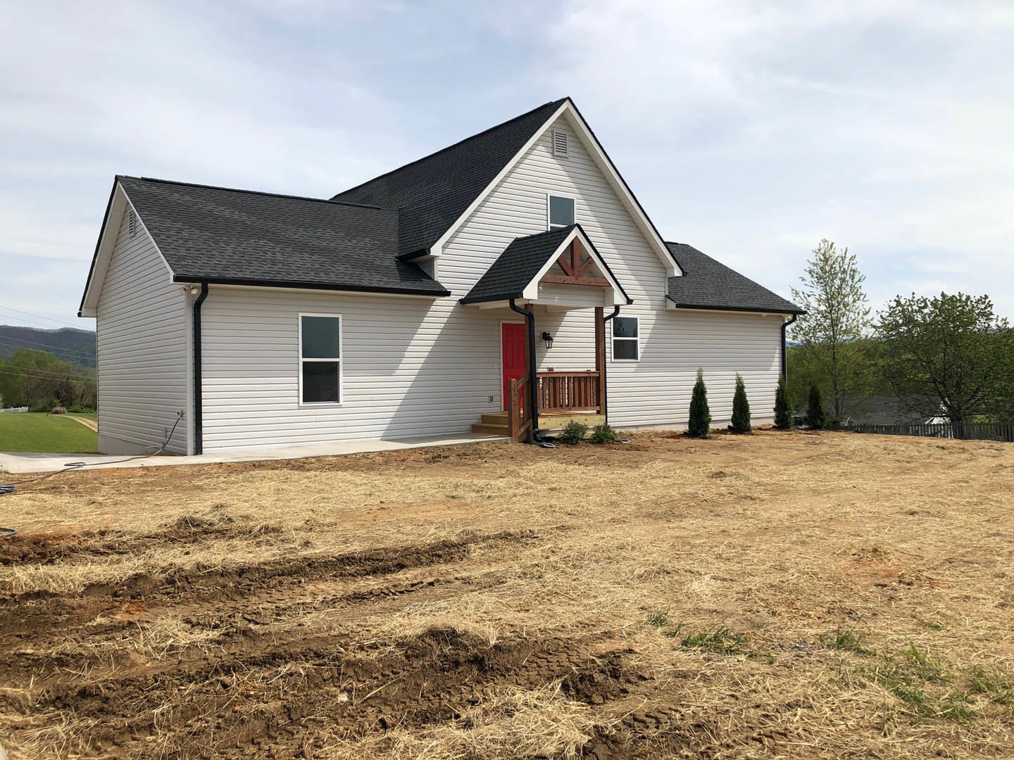 White cottage-style home with red front door, white-framed windows, and covered porch, set beside a large grassy field with a dirt patch and tire tracks; mature tree with green