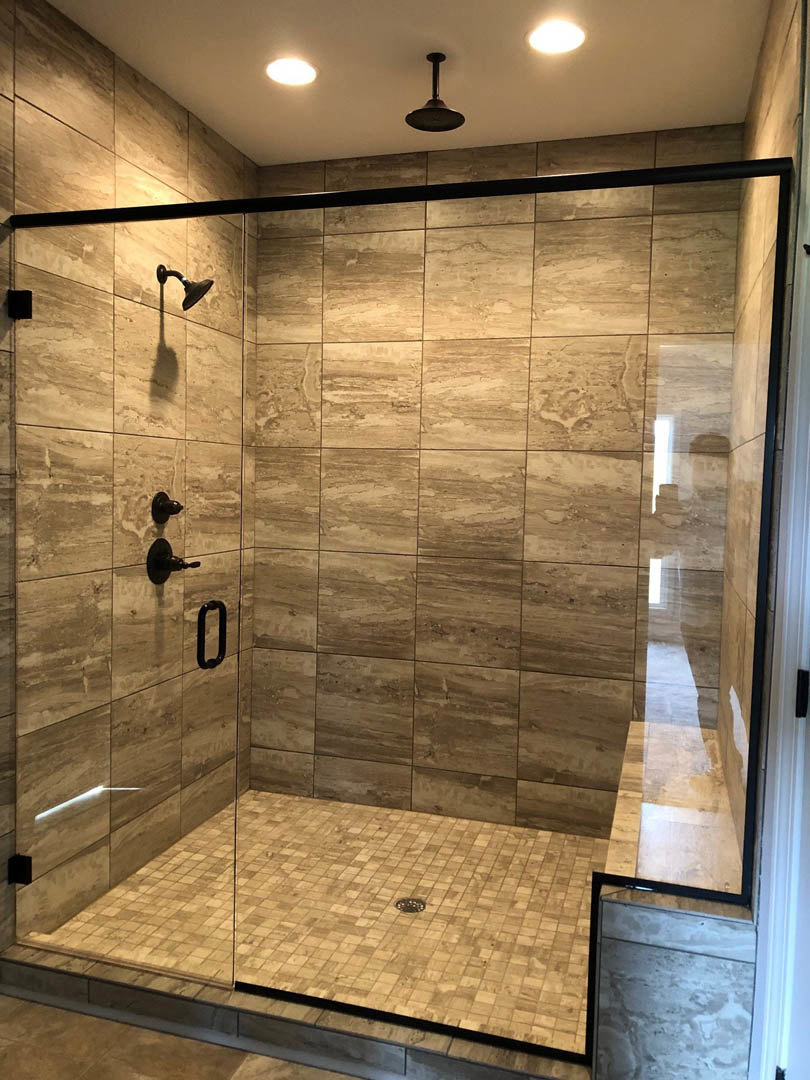 Glass shower enclosure with black handle, white tile walls, round shower head, black ceiling light fixture, and light-colored floor tiles