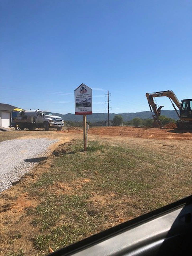 Construction site with grassy field, excavator with extended arm, white utility truck with side tank, and a posted sign on a pole near a white wall with black frame