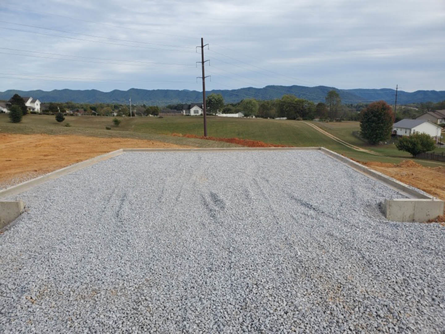 Gravel driveway bordered by green field, leafy tree, and power line pole, with a house partially hidden behind trees and mountains under a cloudy sky in the background