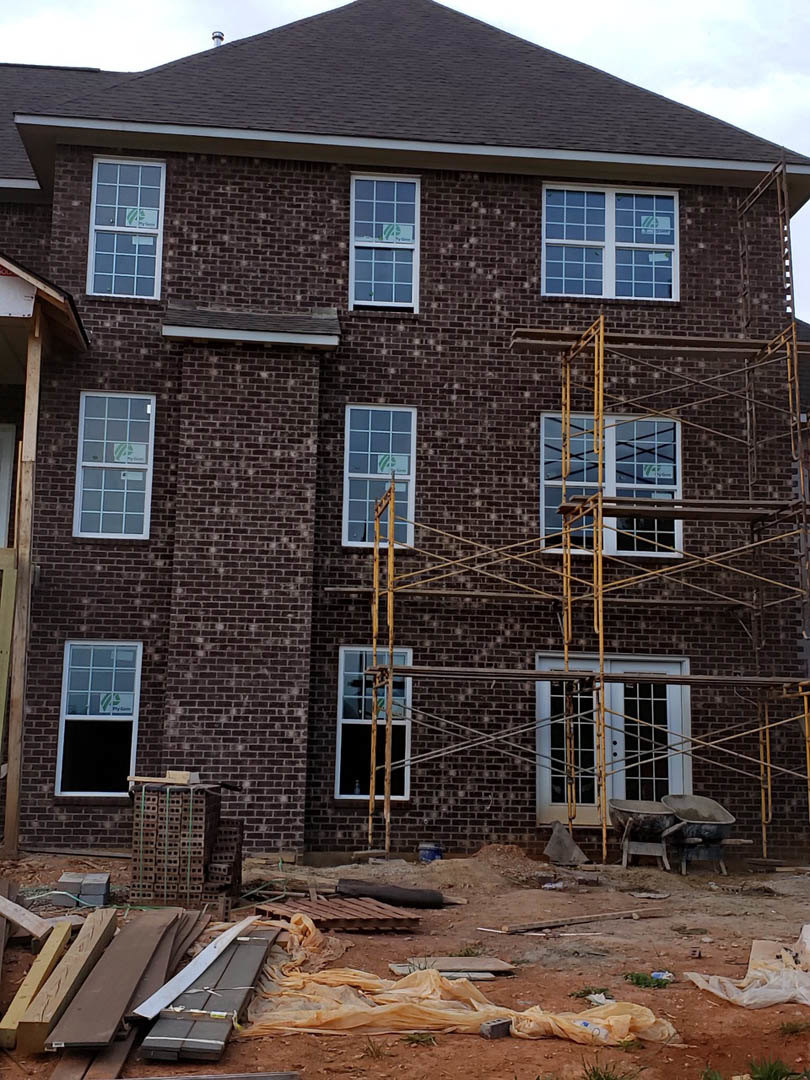 Scaffolding surrounds a partially built house with exposed framing, sash windows, stacked bricks secured by a green strap, and metal strips piled near the foundation.