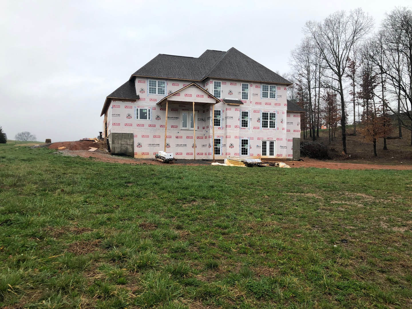 Partially built house with exposed insulation, unfinished roof, several windows, surrounded by grass and mature trees under a cloudy sky