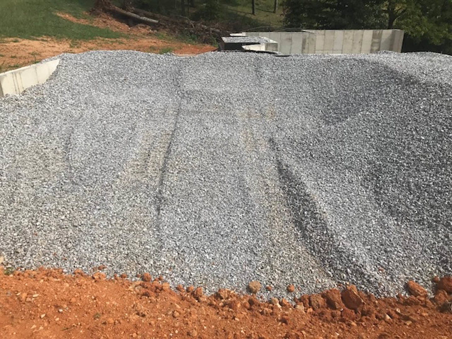 Gravel pile on bare soil beside unfinished foundation wall, construction materials scattered, sparse grass and small tree in background