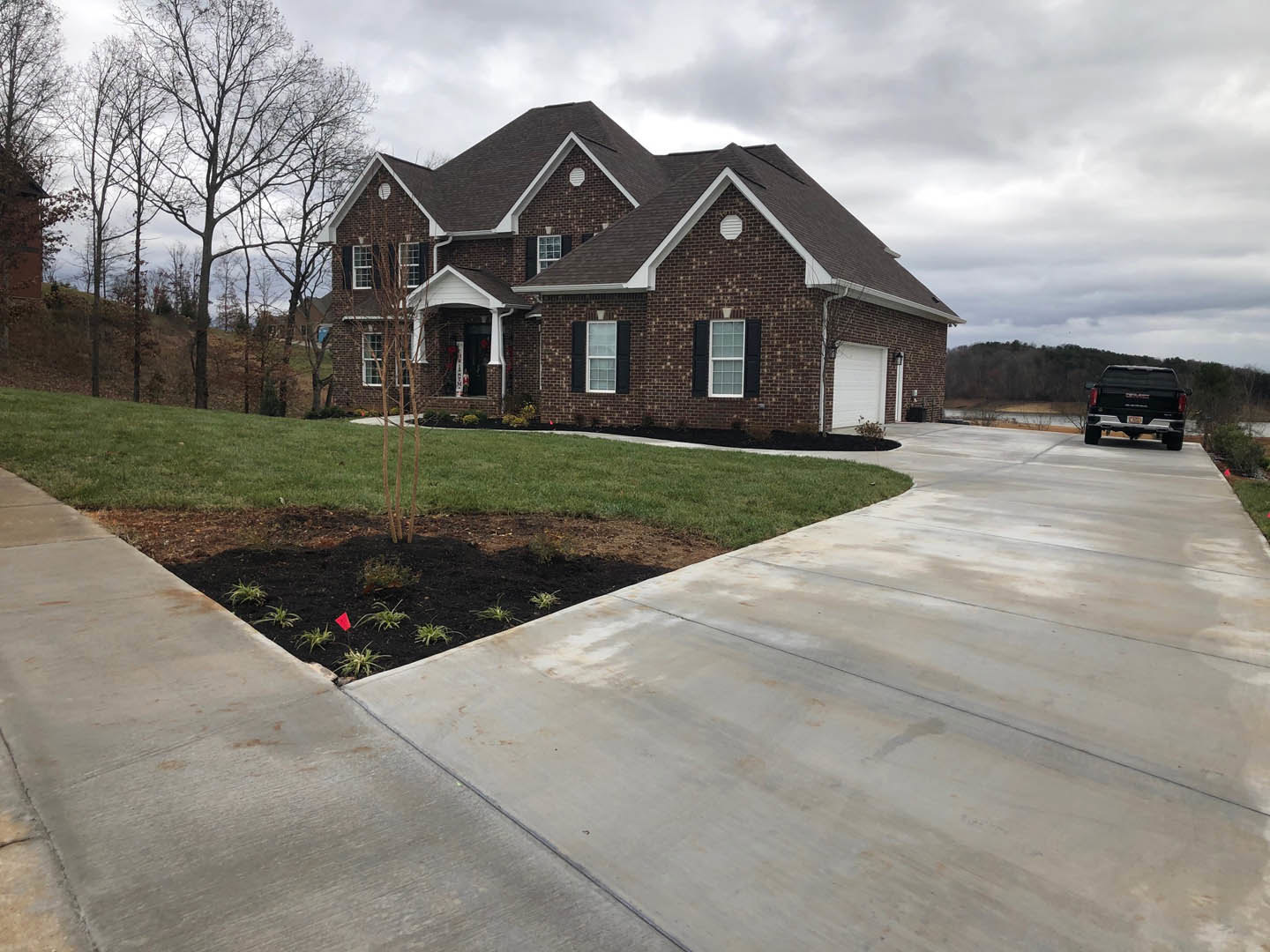 Brick house with black roof, white garage door, concrete driveway, green lawn, leafless tree, and black truck parked on street; sidewalk runs alongside property.